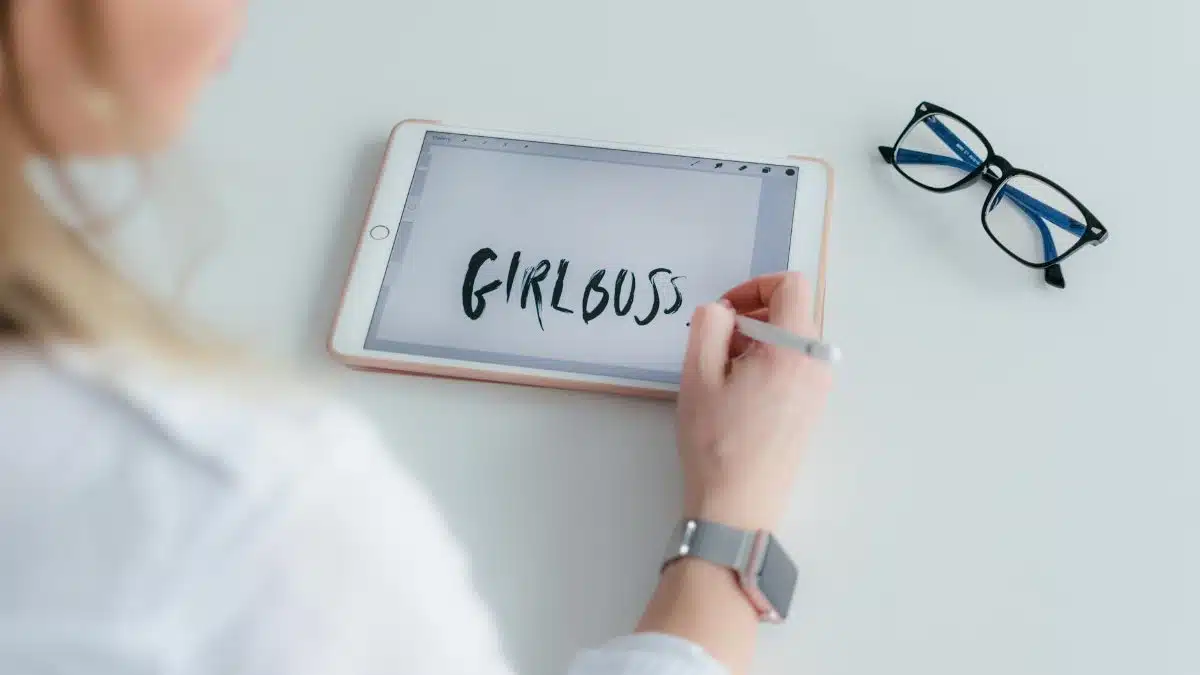 A woman writes 'girlboss' on a tablet while her glasses are placed beside her on a white table.