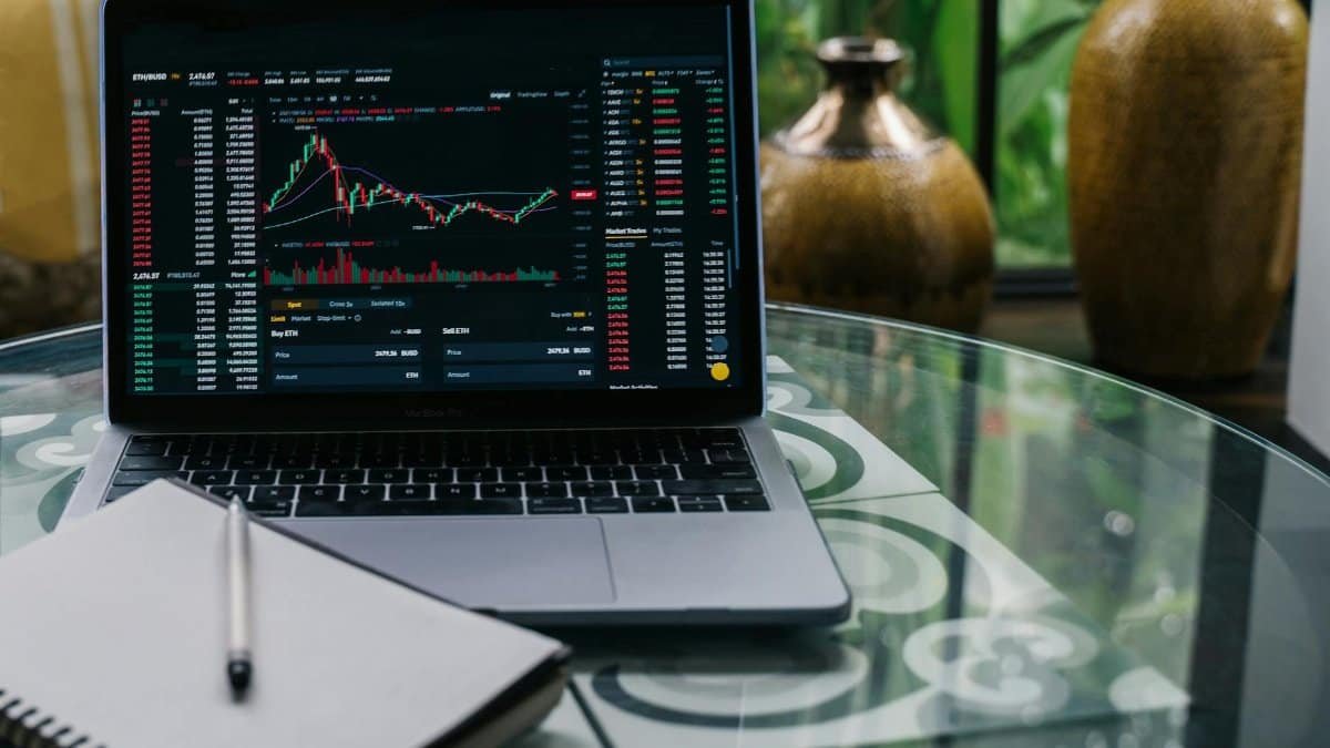 Laptop displaying cryptocurrency stocks and graphs on a glass table with a notepad.