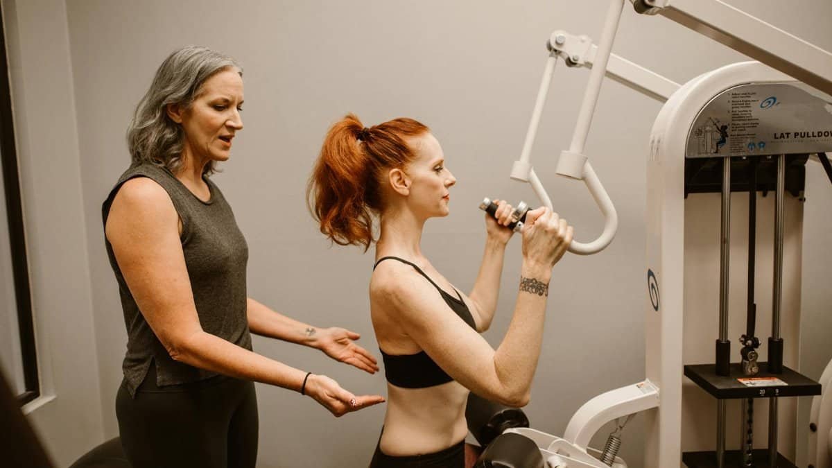 A woman works out on gym equipment with guidance from a trainer indoors.