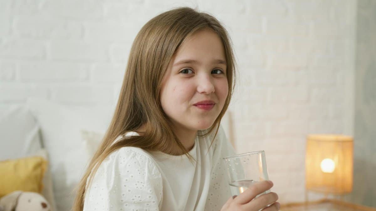 A young girl smiling and holding a glass of water inside a cozy bedroom setting.