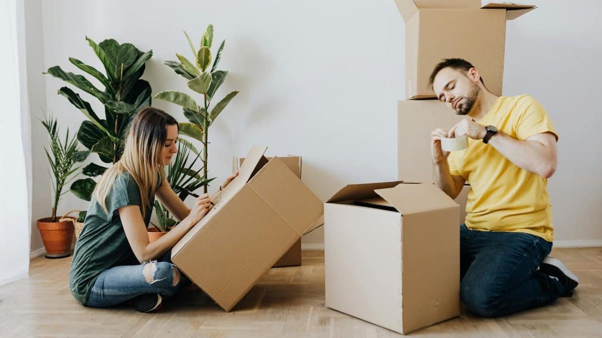 Side view full length glad couple packing stuff into cardboard boxes while sitting on floor at home during relocation in new house