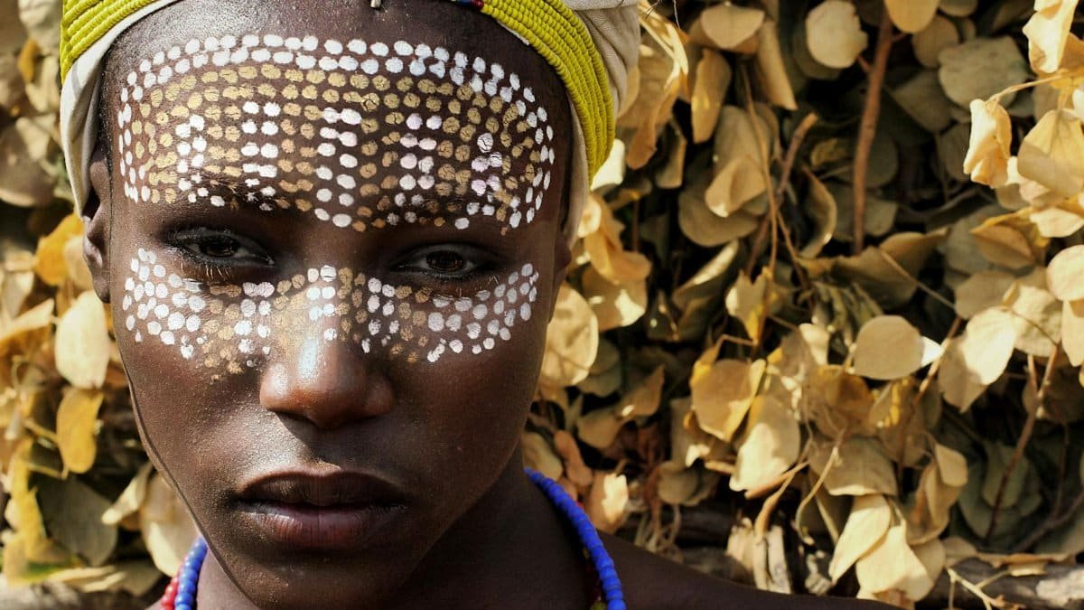 Close-up portrait of an African woman with traditional tribal face paint and beaded adornments.