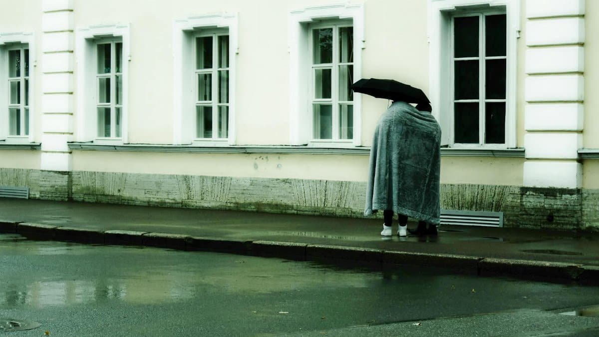 Couple under a blanket sharing an umbrella on an urban sidewalk in rainy Saint Petersburg.