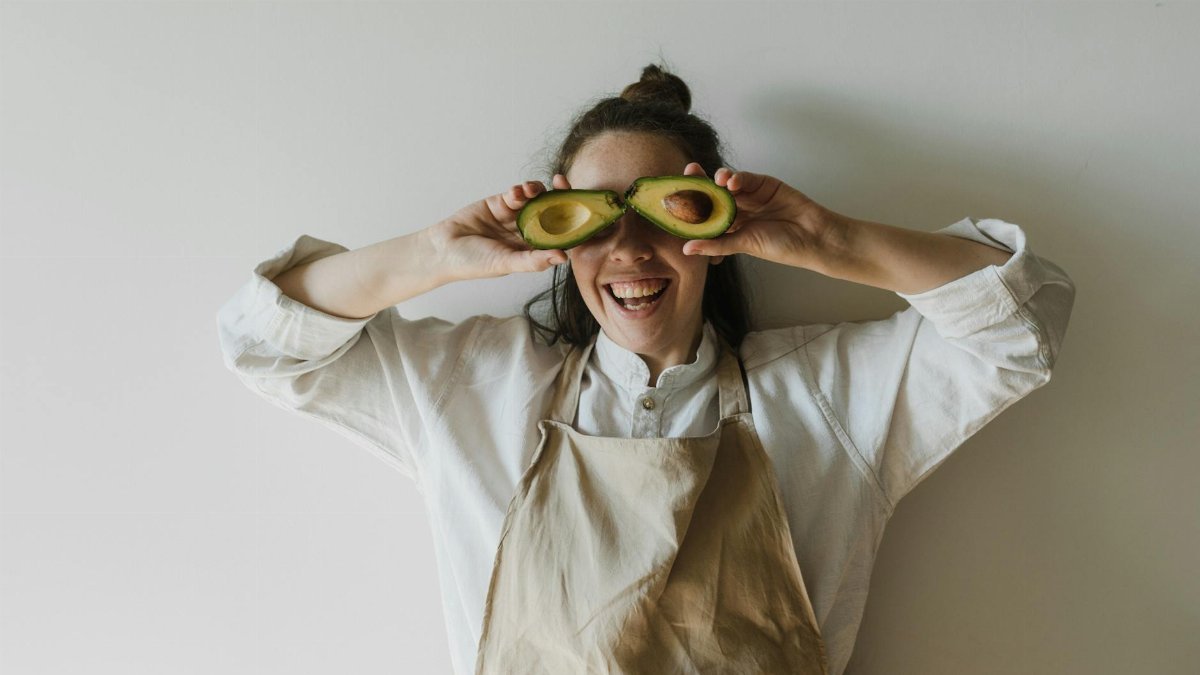 Joyful woman holding avocado halves over her eyes, symbolizing healthy living and fun creativity.