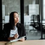 Portrait of a woman in a modern office pondering ideas, holding a notebook and pen.