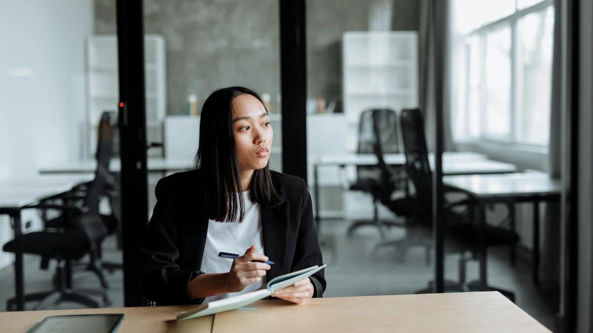 Portrait of a woman in a modern office pondering ideas, holding a notebook and pen.