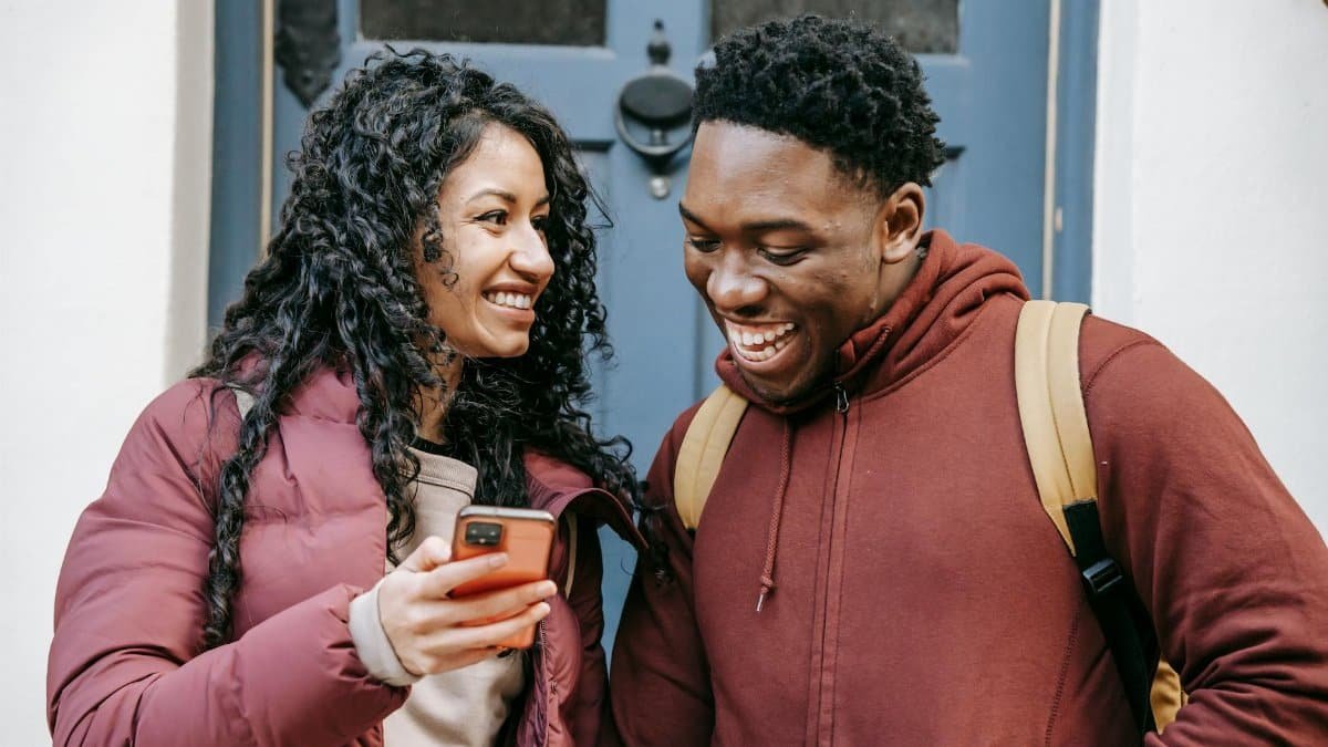 Young happy multiethnic partners talking while browsing internet on cellphone in town in daytime