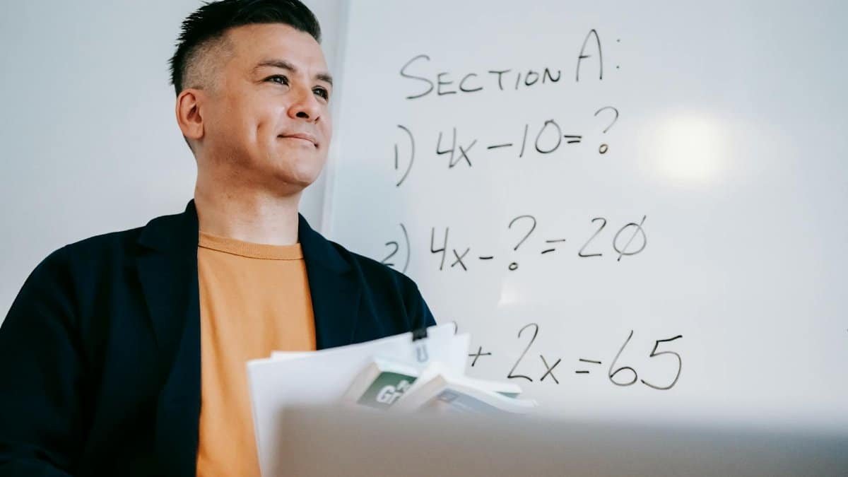 Smiling teacher holding papers, explaining algebra equations on a whiteboard in a classroom setting.