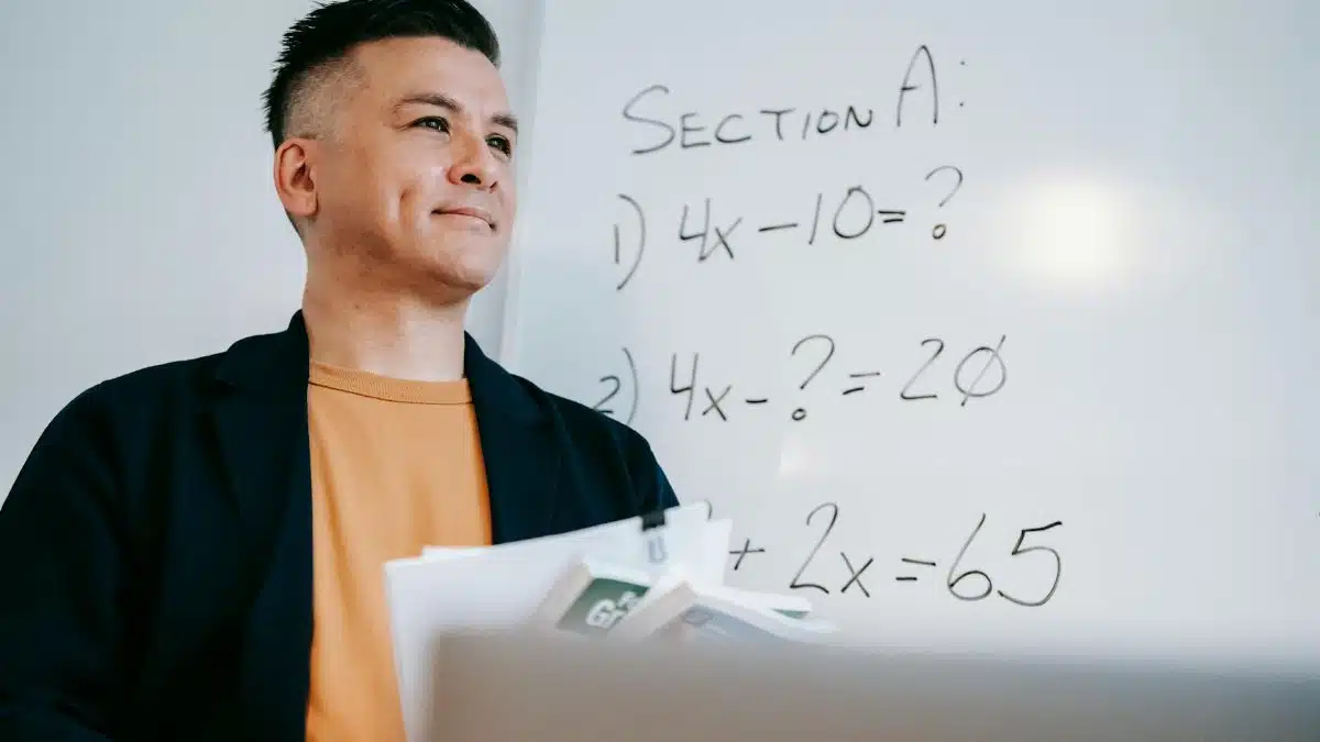 Smiling teacher holding papers, explaining algebra equations on a whiteboard in a classroom setting.