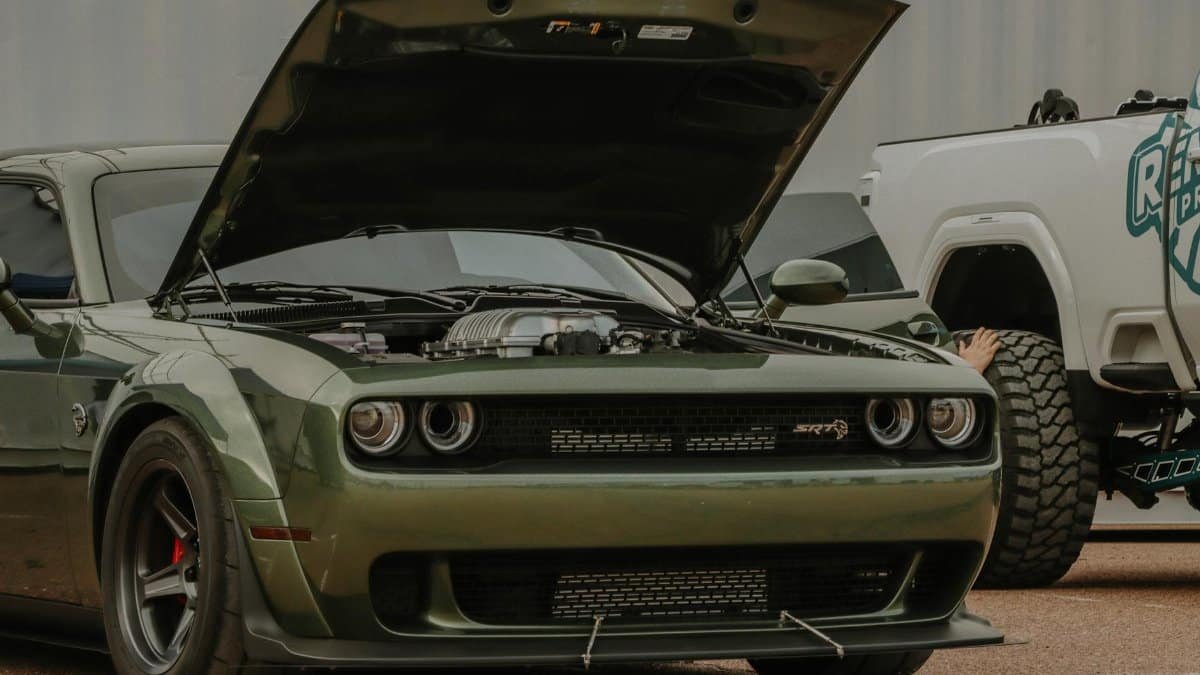 A green Dodge Challenger displayed with its hood raised, showcasing its engine at an outdoor car event.
