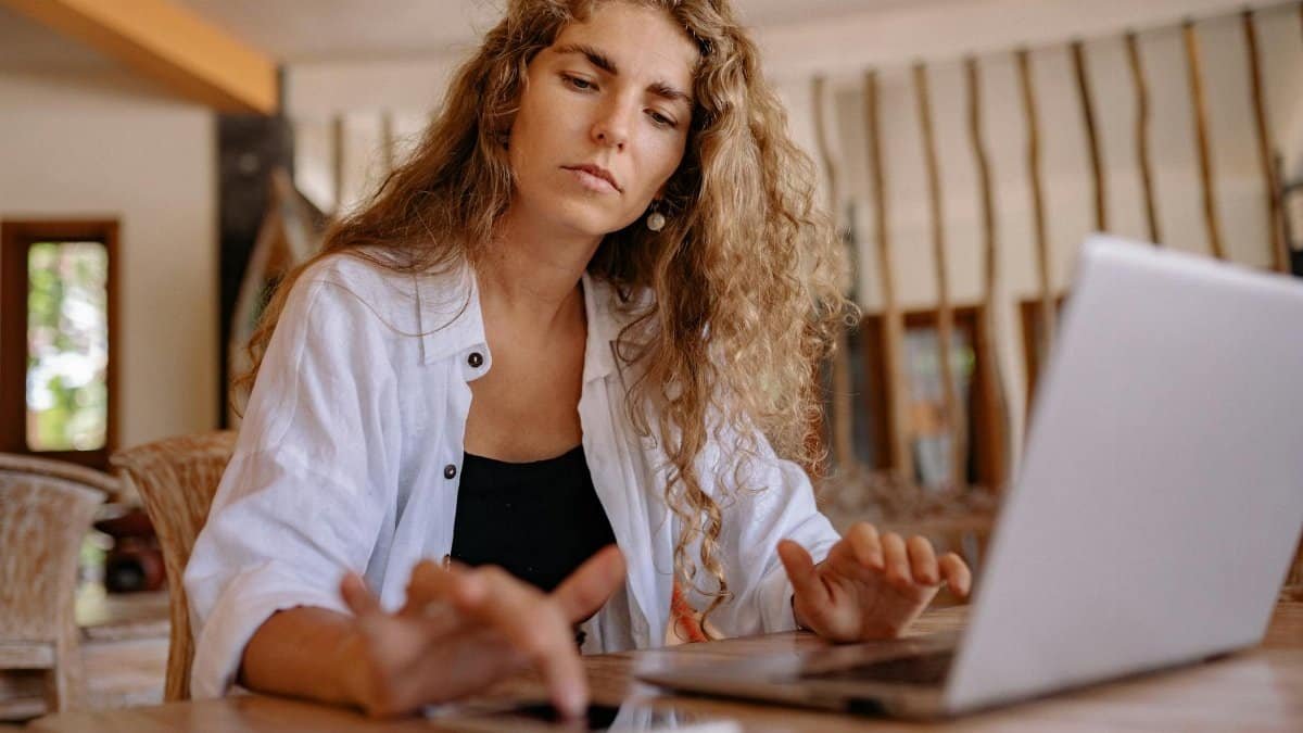 A woman intently works on a laptop while using a smartphone indoors, symbolizing modern remote work.