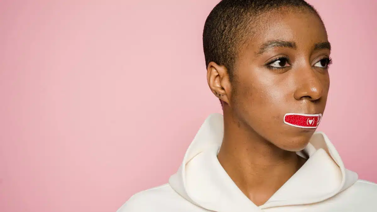 Empowered African American woman with Band-Aid on lips, symbolizing silence and awareness on a pink background.