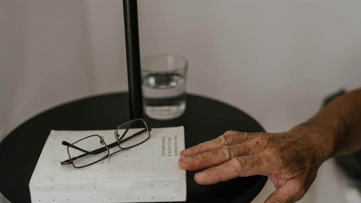 Close-up of an elderly hand reaching for glasses on a bedside table with a book and water glass, in a calm setting.