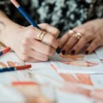 Crop anonymous tattooed female designer coloring apparel illustration on human figure on paper sheet while working at desk