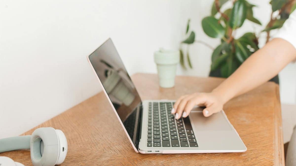 A hand using a laptop on a wooden desk with headphones and a green plant nearby, depicting modern remote work.