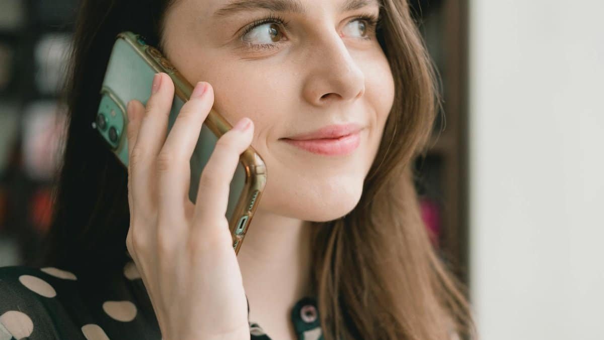 Crop young happy female millennial with long dark hair smiling and looking away while having phone conversation near window on sunny day