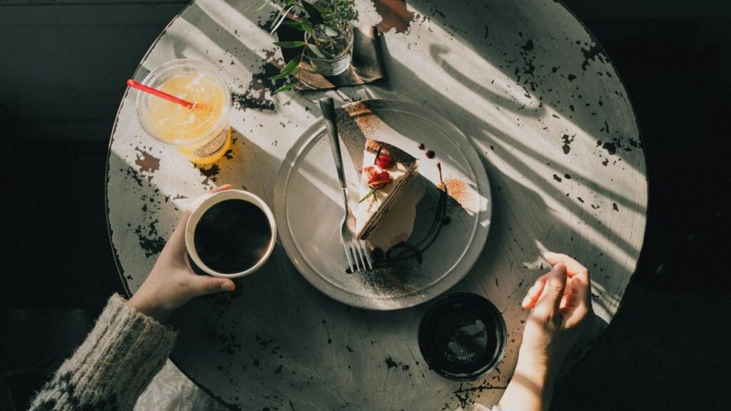 Overhead view of a cozy café table with a slice of cake, coffee, and juice, bathed in warm afternoon light.