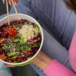 Vibrant poke bowl with vegetables and grains, held in hands, viewed from above.