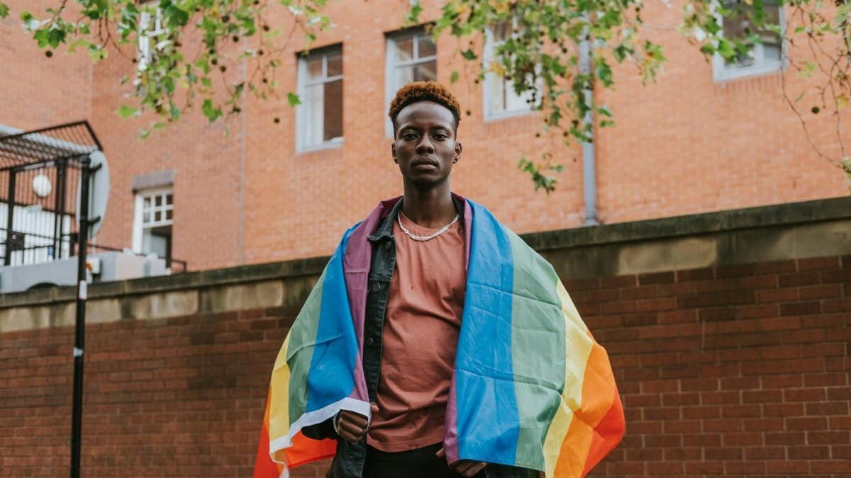 Serious African American male with LGBT flag looking at camera while standing on street against brick building on street