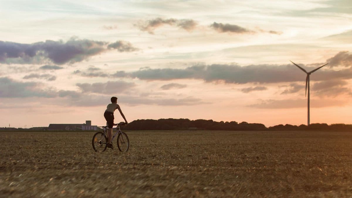 Cyclist rides through field with wind turbine at sunset, symbolizing renewable energy.