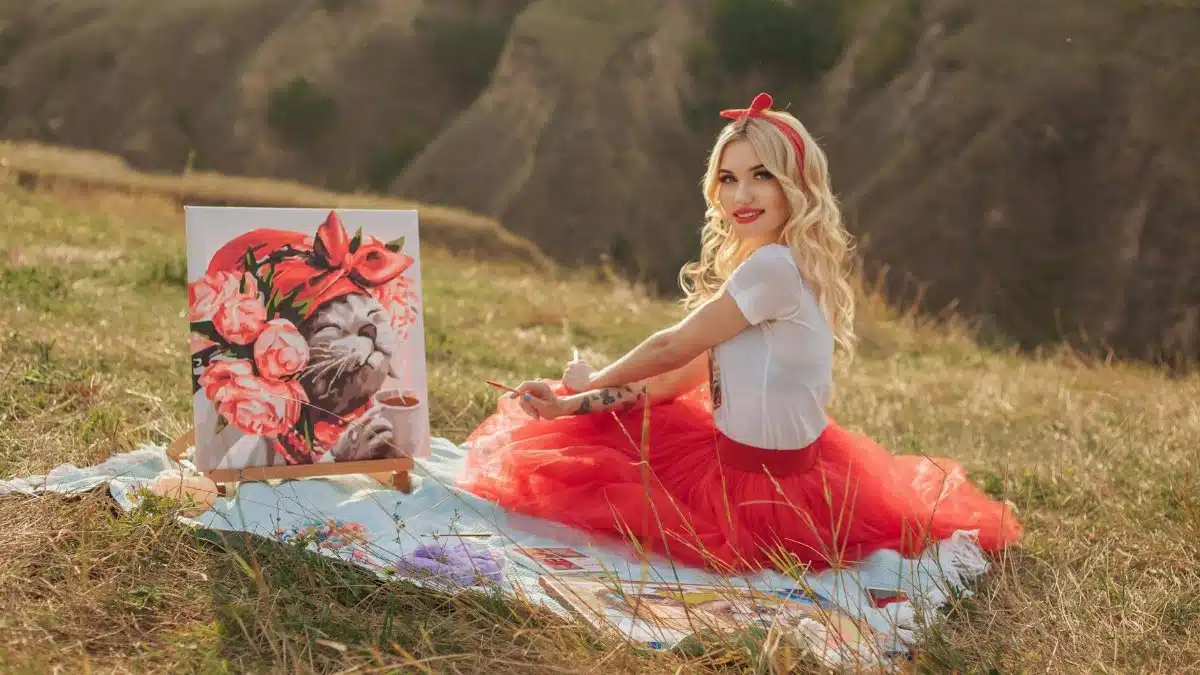 A smiling woman in a red tutu skirt paints in a sunny London meadow.