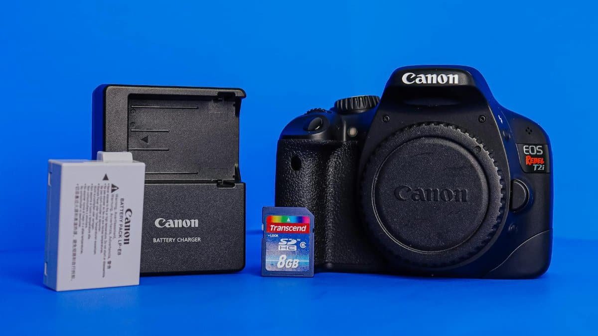 A Canon DSLR camera accompanied by a battery, charger, and memory card against a blue background.