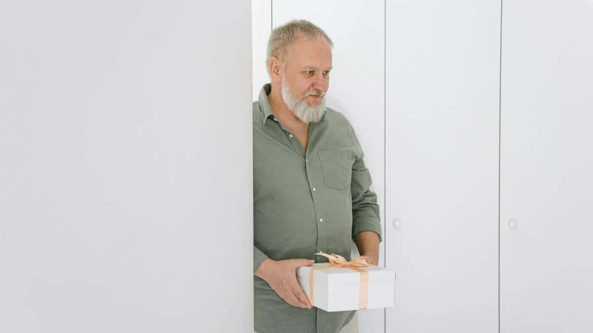 Elderly man in a green shirt holding a gift box in a bright, minimalist room.