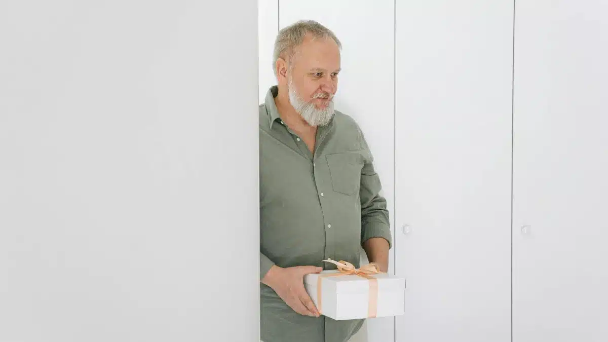 Elderly man in a green shirt holding a gift box in a bright, minimalist room.