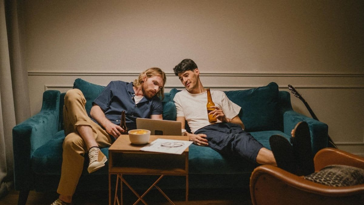 Two friends sitting on a couch, enjoying drinks and watching a laptop in a cozy living room.