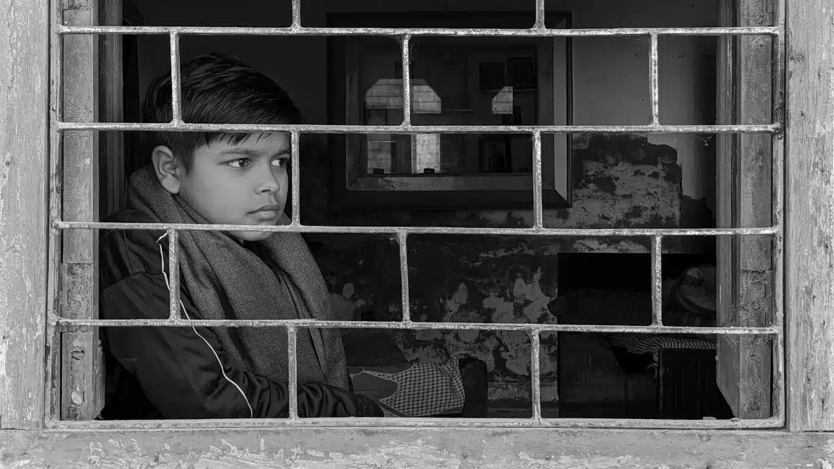 A young boy gazes outside through barred windows, symbolizing isolation.
