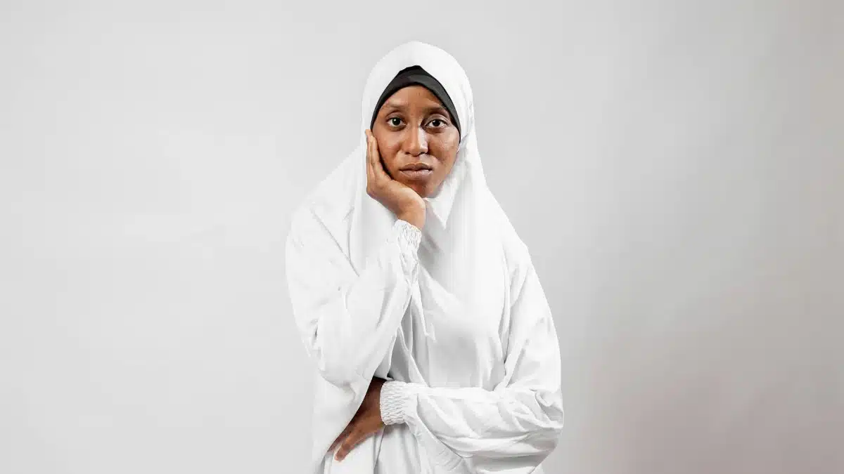 A portrait of a thoughtful woman wearing a traditional white hijab against a plain background.