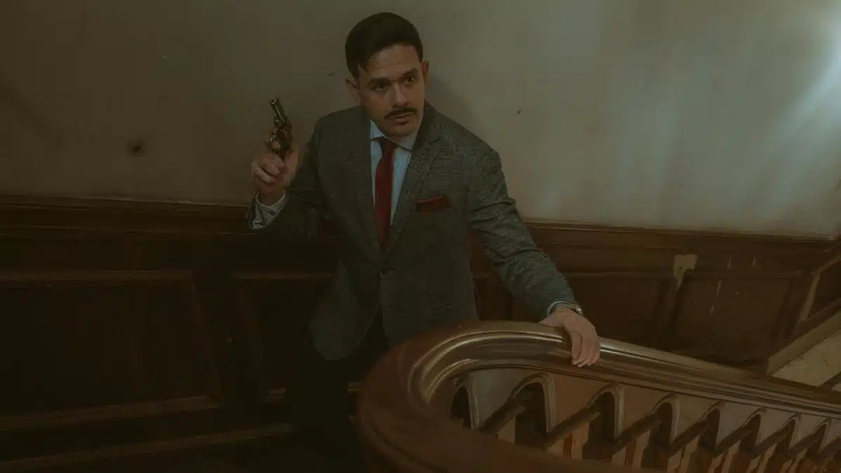 Moody scene of a man in a suit with a revolver on an ornate staircase.