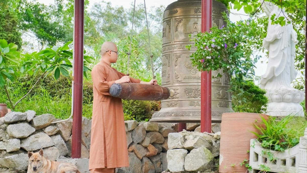A Buddhist monk stands next to a large bronze bell in a temple garden in Thạch Lâm, Việt Nam.