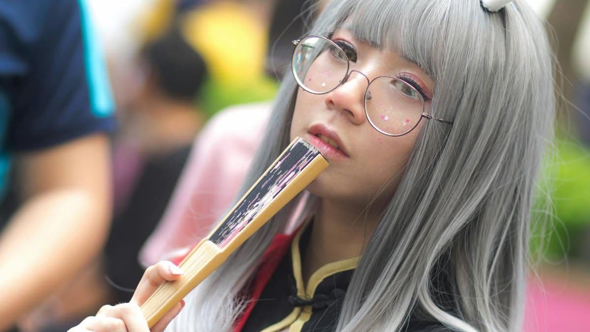 A stylish young woman in cosplay, wearing glasses and holding a fan, posing outdoors.