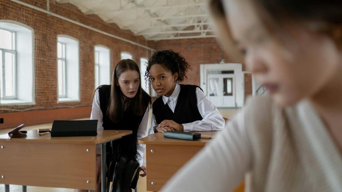 A group of teenagers whispering in a classroom setting with desks and exposed brick walls.