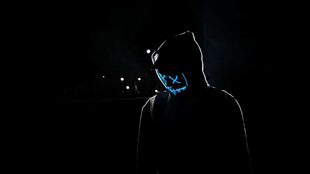 Silhouette of a person in a hoodie wearing a glowing LED mask against a dark background.