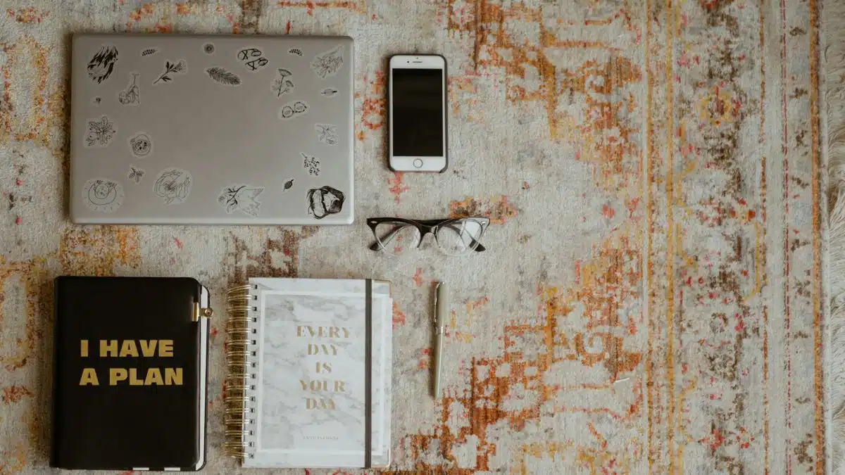 Flat lay of workspace items including laptop, phone, planners, and glasses on a patterned rug.