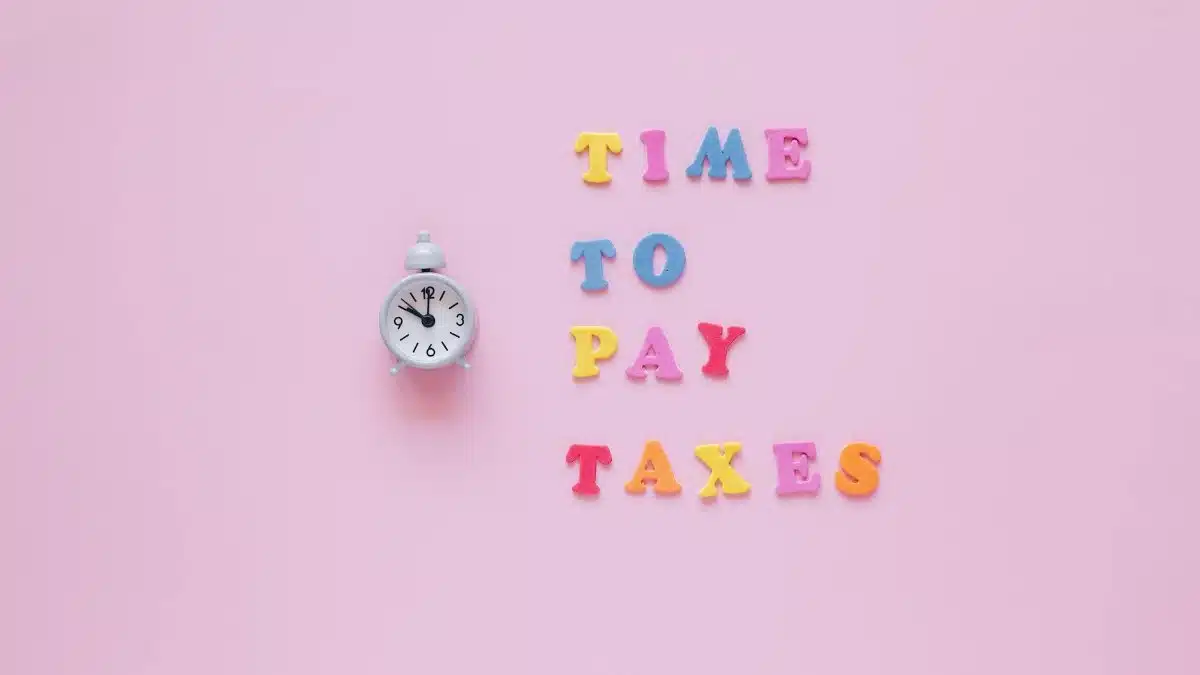 Colorful letters spell 'Time to Pay Taxes' next to a clock on a pink surface.