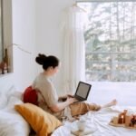 Side view of cute toddler girl sitting on bed barefoot and playing with colorful wooden blocks while mother using laptop in bed enjoying morning coffee