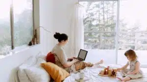 Side view of cute toddler girl sitting on bed barefoot and playing with colorful wooden blocks while mother using laptop in bed enjoying morning coffee