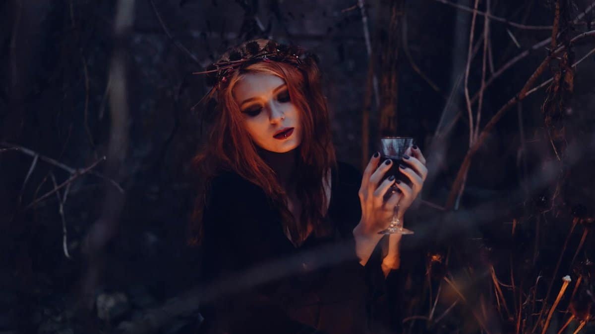A mystical woman in gothic attire holds a chalice amidst a dark, eerie forest setting.
