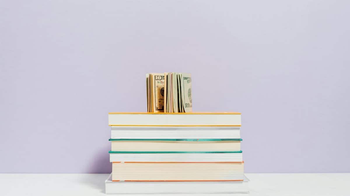 Stack of books with a stack of US dollar bills on top against a pastel background.