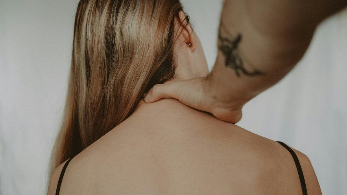 Anonymous abusive man with tattoo touching neck of vulnerable woman sitting on white background during domestic violence in light room