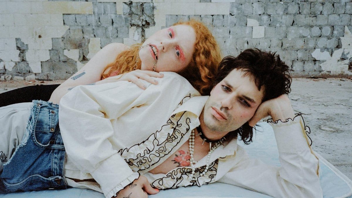 A couple in avant-garde fashion poses against a distressed wall, showcasing unique style.