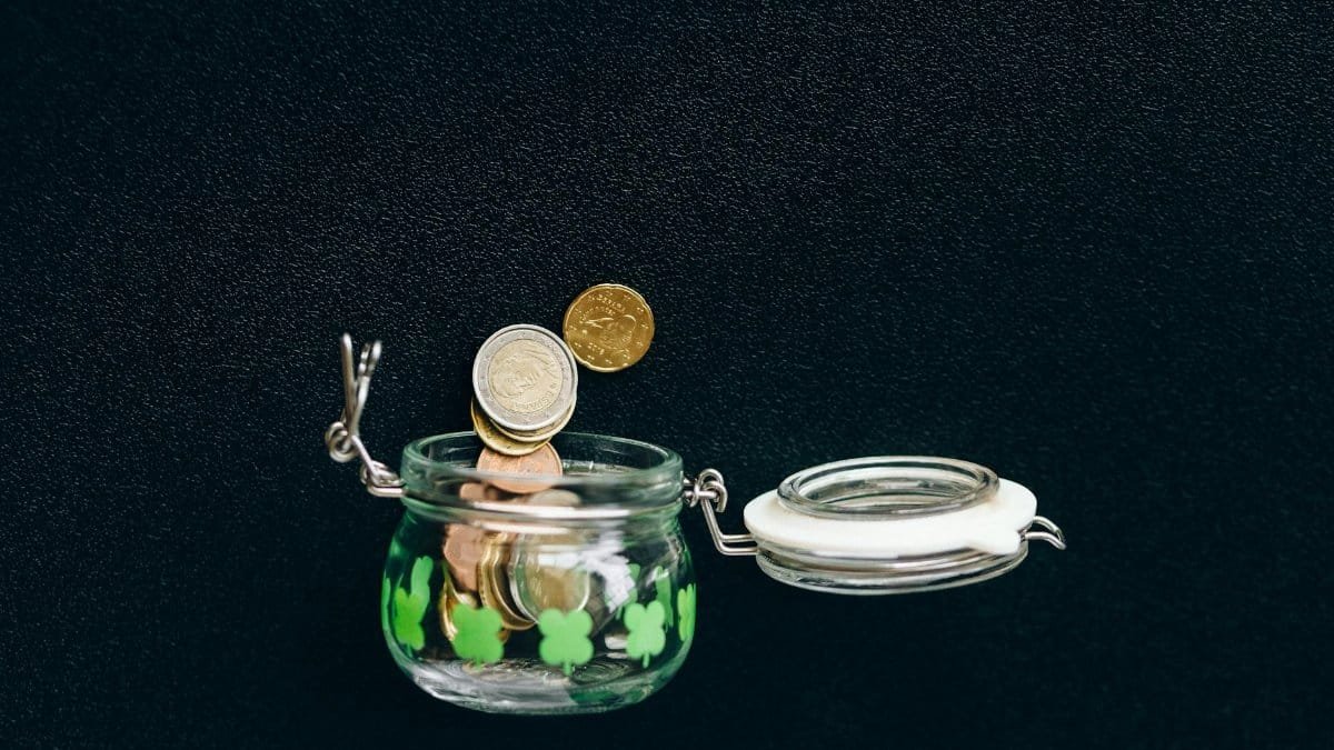 Glass jar with coins falling into it on a black background, symbolizing savings.
