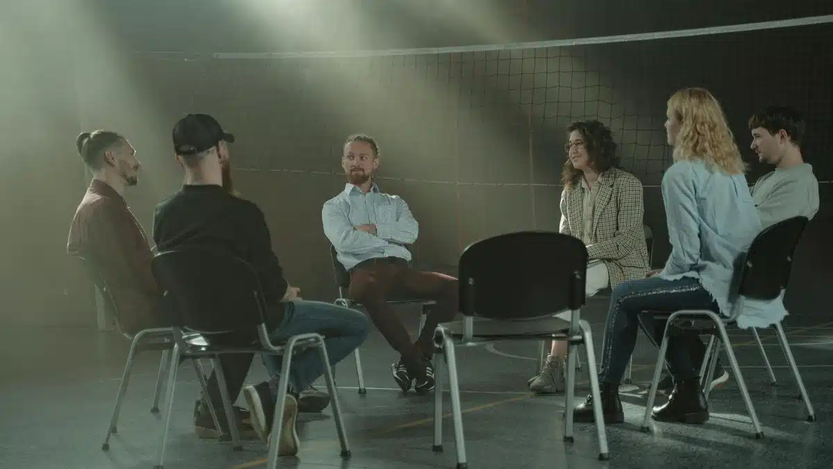 A group therapy session with six adults seated in a circle, discussing support and mental health.