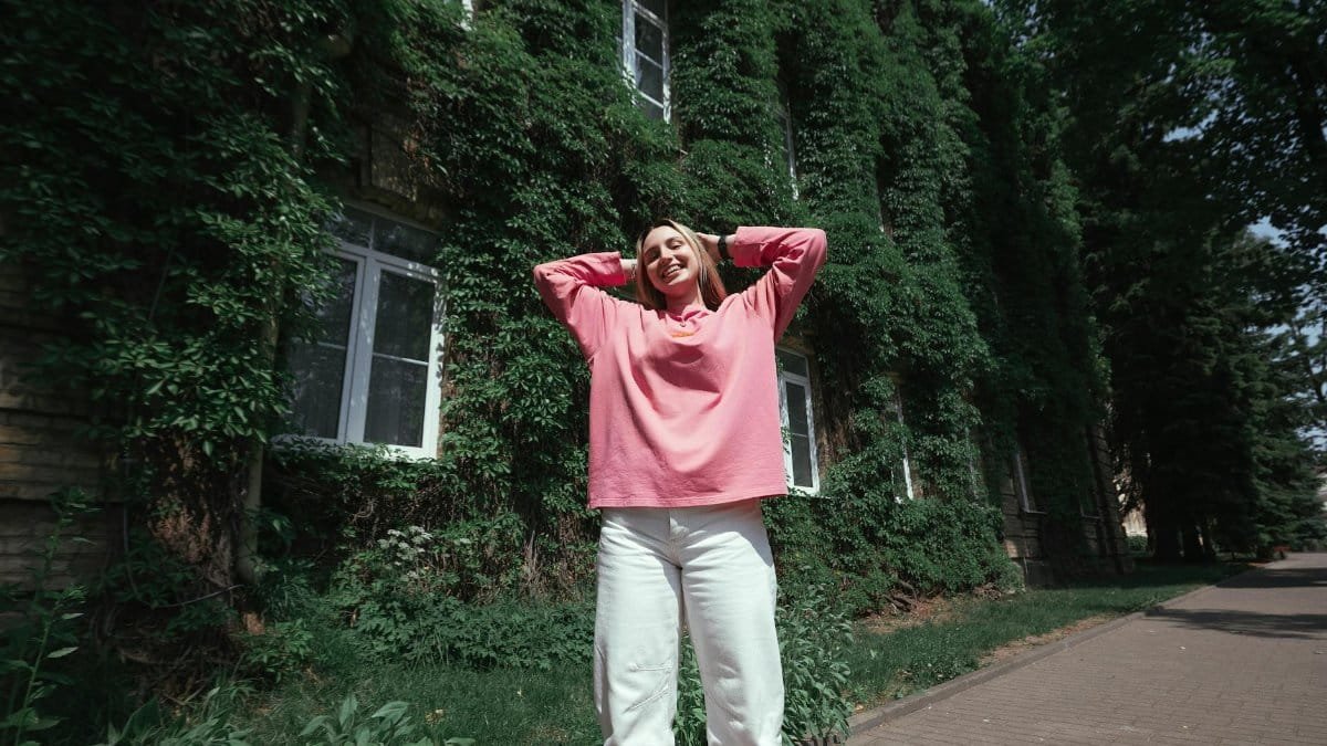 Smiling young woman in pink enjoying a sunny day outdoors with ivy-covered building.