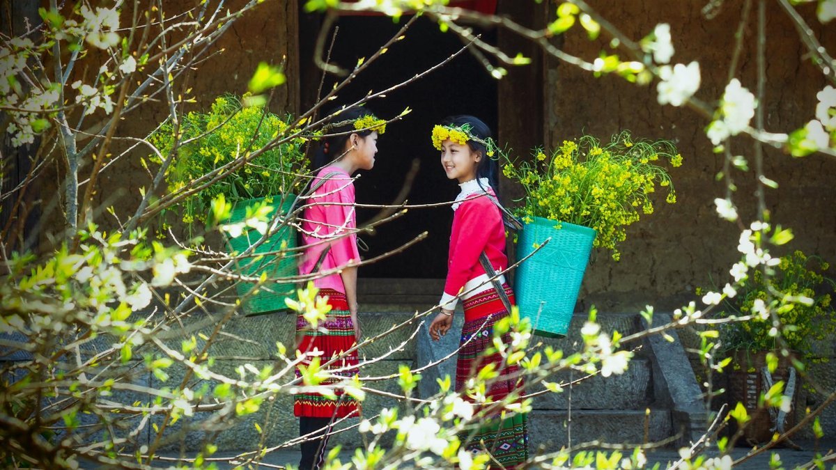 Two girls in traditional dress carrying flowers outdoors, surrounded by spring foliage.