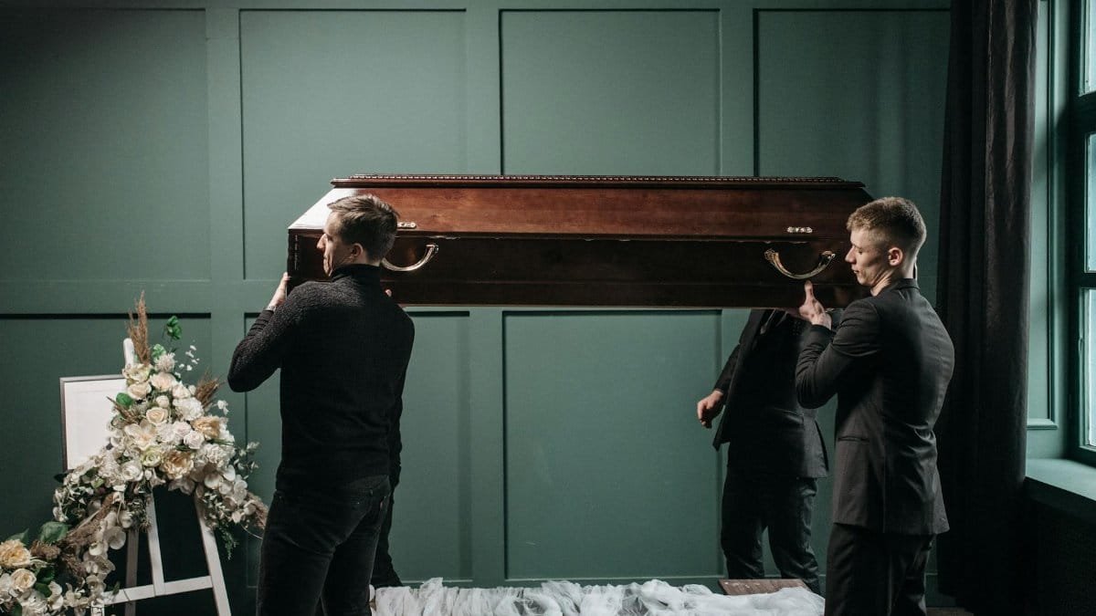 Pallbearers carry a wooden coffin at an indoors funeral service, surrounded by floral tributes.