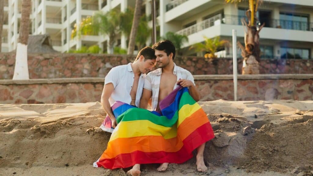 Two men sitting closely on a beach, wrapped in a vibrant rainbow flag, showcasing warmth and intimacy.